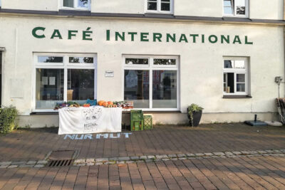 Hausfront mit dem Café International, vor den Fenstern des Cafés ein Tisch mit Gemüse und einem Banner "Regionalkollektiv"